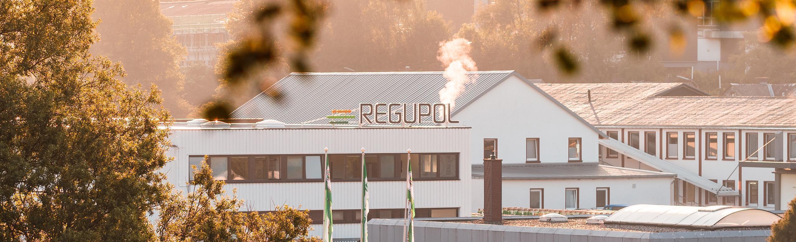 REGUPOL Produktionsstandort im Morgenlicht – Werk, Nachhaltigkeit, Industrie Außenansicht eines REGUPOL-Produktionsstandorts in morgendlichem Licht. Das weiße Industriegebäude trägt das REGUPOL-Logo auf dem Dach, aus einem Kamin steigt Dampf. Im Vordergrund sind Bäume und mehrere grüne REGUPOL-Fahnen zu sehen.